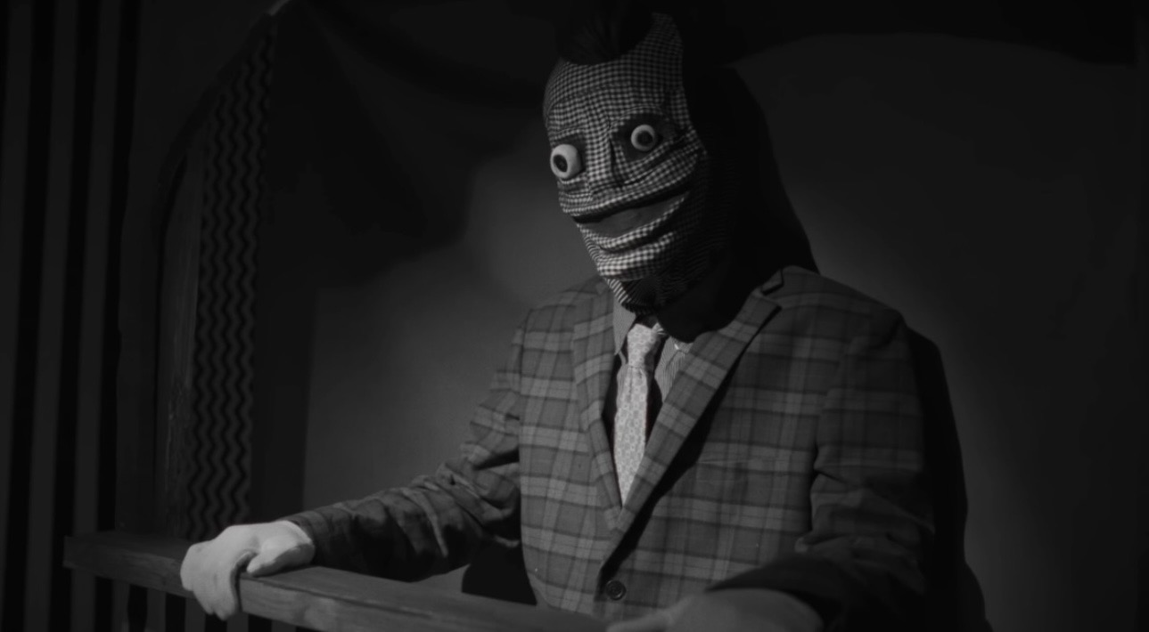 Black-and-white frame of a masked figure in a checked suit
